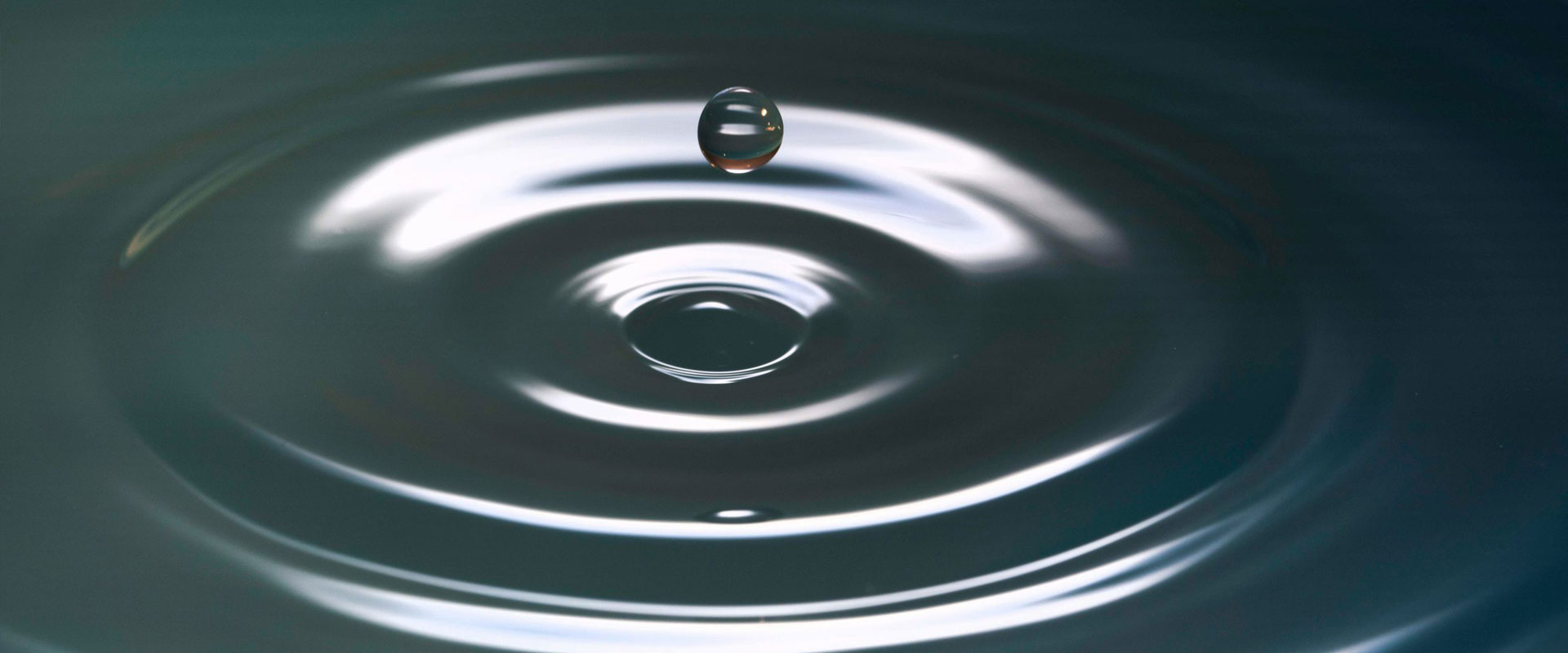 Wassertropfen schwebt über einer ruhigen Oberfläche und erzeugt konzentrische Wellen.