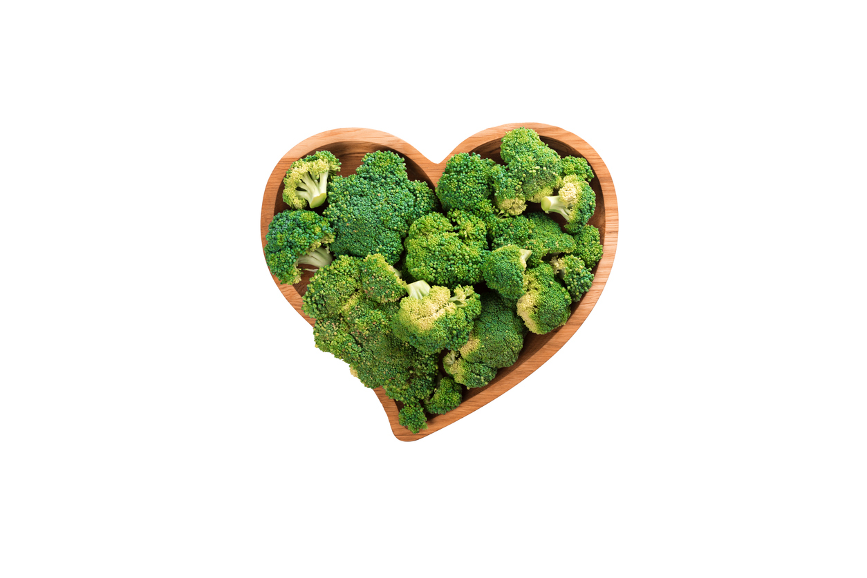 Green broccoli florets fill a heart-shaped wooden bowl, centered on a plain white background.